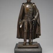 Escultura em bronze. Figura masculina, voltada para frente, cabeça com cabelos, face com  costeleta, veste farda militar e capa sobre os ombros. Na mão direita segura um chapéu e a mão esquerda repousa sobre a espada presa à cintura.